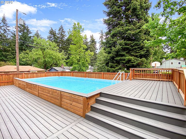 The gas-heated 32-by-16-foot above-ground pool surrounded by a large Trex deck, with the wooden privacy fence and tall firs behind